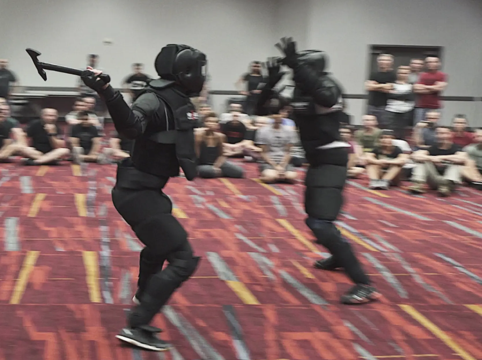 Two students in HIGH GEAR armour running full-contact force-on-force scenarios in front of a seminar class